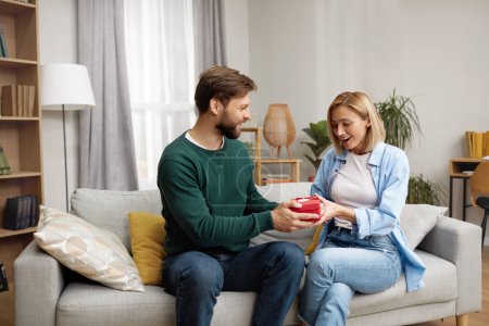 Happy Man Presenting Gift To Wife. Excited Young Man Presenting Good Unexpected Present To Wife At Home. Loving Boyfriend Making Romantic Surprise To Girlfriend 
