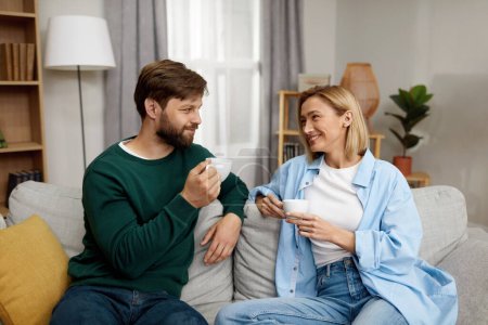 Téléchargez les photos : Happy Couple Drinking Coffee At Home. Young Couple Talking And Holding Cups While Sitting On Beige Sofa At Home. Domestic Lifestyle Concept - en image libre de droit