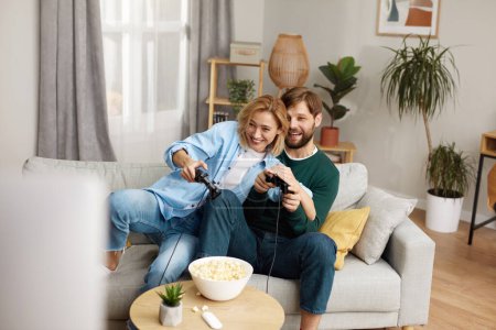 Foto de Young Couple Playing Video Games. Boyfriend And Girlfriend Sitting On Couch In Living Room Enjoying Playing Video Games And Spending Time Together. Enjoying Moment On Weekend Concept - Imagen libre de derechos