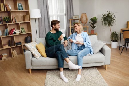 Téléchargez les photos : Happy Couple Drinking Coffee At Home. Young Couple Talking And Holding Cups While Sitting On Beige Sofa At Home. Domestic Lifestyle Concept - en image libre de droit