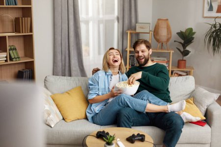 Téléchargez les photos : Laughing Couple Watching TV At Home. Loving People Resting On Sofa And Eating Popcorn At Cozy Home Morning. Leisure Concept - en image libre de droit