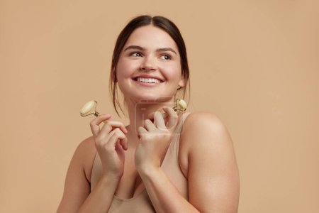 Photo for Face massage. Plus Size Woman Holding Jade Facial Roller for Skin Care, Beauty Treatment on Beige Isolate Background. Girl Taking Care of Skin with Natural Massager Closeup Portrait - Royalty Free Image