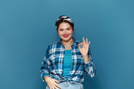 Photo for Smiling Woman Ok Gesture Isolated. Portrait of Satisfied Plus Size Girl Standing, Looking at Camera and Showing Ok Sign Gesture. Indoor Studio Shot Isolated on Blue Background - Royalty Free Image