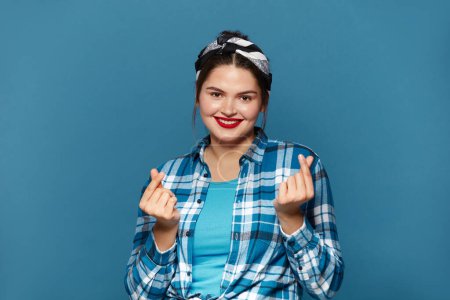 Photo for Smiling Woman Showing Money Gesture. Portrait of Plus Size Girl Smiling and Showing Give Me Money Gesture, Asking for Payment. Indoor Studio Shot Isolated on Blue Background - Royalty Free Image