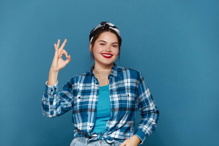 Téléchargez les photos : Smiling Woman Ok Gesture Isolated. Portrait of Satisfied Plus Size Girl Standing, Looking at Camera and Showing Ok Sign Gesture. Indoor Studio Shot Isolated on Blue Background - en image libre de droit