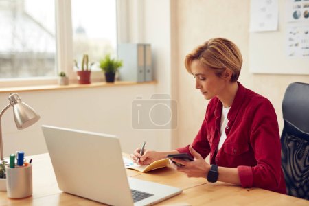 Photo for Business Woman Working At Office. Focused Lady Using Laptop Writing Notes. Female Person Holding Mobile Sitting At Workplace In Modern Office. Successful Entrepreneurship And Career Concept - Royalty Free Image