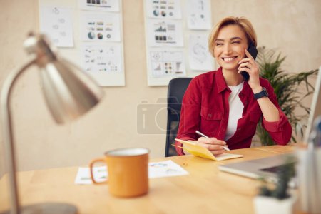 Smiling Businesswoman Calling Smartphone. Positive Blonde Woman Talking On Phone in Office Alone. Happy Lady Writing Notes Holding Mobile. Business concept 