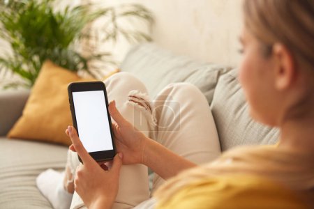 Foto de Woman Using Smartphone At Home. Blonde Girl Sitting On Sofa And Messaging In Cozy Living Room at Home. Female Person Doing Online Shopping, Browsing Internet and Checking Social Media - Imagen libre de derechos