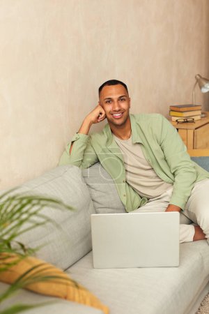 Photo for Smiling Man Using Laptop At Sofa. Young Guy Holding Computer, Sitting At Home On Cozy Couch. Multiracial Man Relaxing With Laptop And Looking At Camera - Royalty Free Image