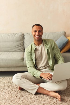 Photo for Smiling Man Using Laptop At Home. Young Guy Holding Computer, Sitting At Floor At Cozy Home. Multiracial Man Relaxing With Laptop And Looking At Camera - Royalty Free Image