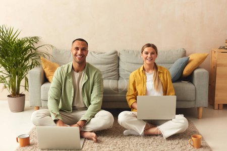 Photo for Couple Using Laptops At Home. Married Couple Of Freelancers Work From Home. Guy And Girl In Casual Clothes With Gadgets Work In The Living Room At The Floor - Royalty Free Image