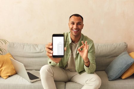Téléchargez les photos : Man Showing Smartphone At Home. Handsome Multiracial Guy Sitting On Sofa And Demonstrating Mobile With Blank White Screen In Cozy Living Room at Home - en image libre de droit
