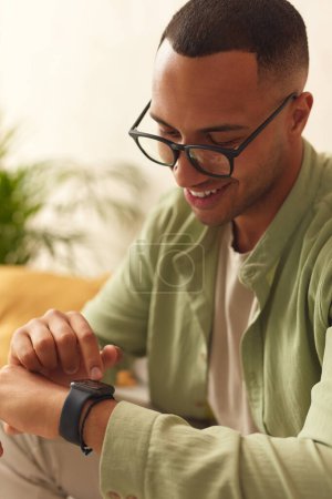Téléchargez les photos : Smiling Man Using Smart Watch. Multiracial Guy Touching Wrist Watch While Sitting Sofa In Living Room. Positive Person Trying New Technologies - en image libre de droit