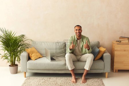 Téléchargez les photos : Happy Man Showing Like Gesture. Optimistic Handsome Guy Showing Thumbs Up Gesture And Smiling, While Sitting At Sofa, Meaning Well Done, Good Job - en image libre de droit