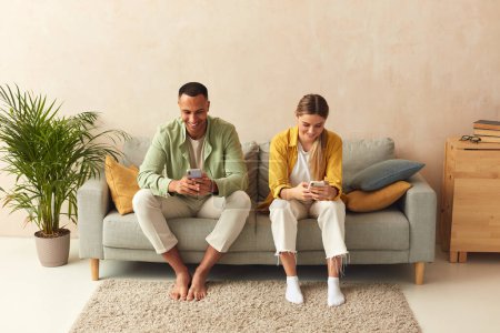 Photo for Couple Messaging Smartphones At Home. Married Couple Relaxing With Mobiles At Home. Guy And Girl In Casual Clothes With Gadgets Sitting In The Living Room At The Sofa - Royalty Free Image