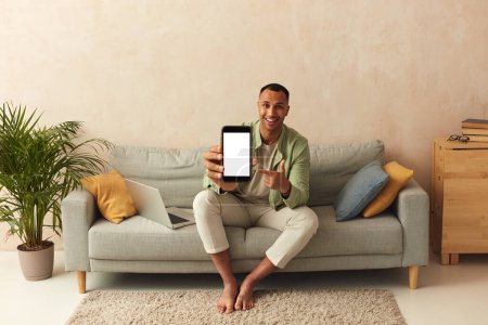 Téléchargez les photos : Man Showing Smartphone At Home. Handsome Multiracial Guy Sitting On Sofa And Demonstrating Mobile With Blank White Screen In Cozy Living Room at Home - en image libre de droit