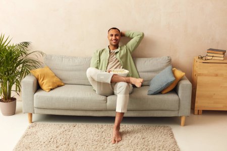 Happy Man Relaxing Sofa. Smiling Middle Eastern Guy Sitting On Comfortable Couch And Eating Pop Corn At Home In Living Room. Cheerful Man Relaxing On Sofa, Enjoying Weekend