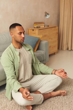 Téléchargez les photos : Calm Man Meditating Home. Multiracial Guy Sitting In Lotus Pose Practicing Yoga At Home. Sport, Fitness And Healthy Lifestyle Concept - en image libre de droit