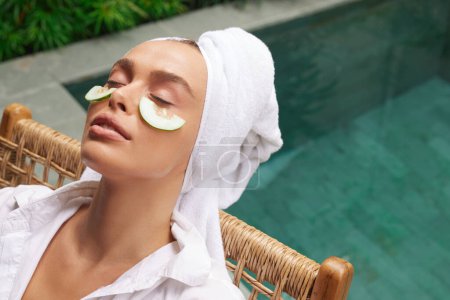 Téléchargez les photos : Relaxed Woman With Cucumber Mask. Young Bathrobe Girl Relaxing With Cucumber Slices Under Eyes Near Swimming Pool. Concept of Skincare - en image libre de droit