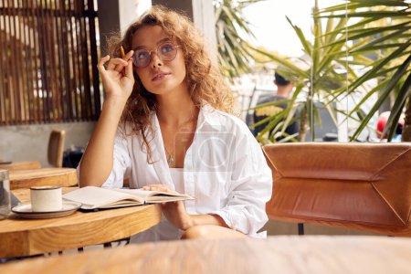 Foto de Freelancer At Cafe. Thoughtful Woman Girl In Glasses Holding Pen While Working In Restaurants. Remote Job Or Education With Modern Digital Technologies For Comfortable Lifestyle In City - Imagen libre de derechos