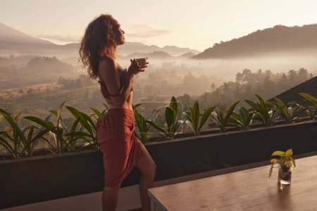 Foto de La mujer disfruta de un tranquilo retiro al amanecer con una taza de café, con vistas impresionantes en Bali. Conceptos de viaje y relajación. - Imagen libre de derechos