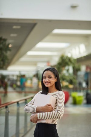 Téléchargez les photos : Une femme adulte joyeuse debout dans un centre commercial, souriant et profitant d'une séance de shopping pendant le week-end. Elle tient la boîte et semble heureuse - en image libre de droit