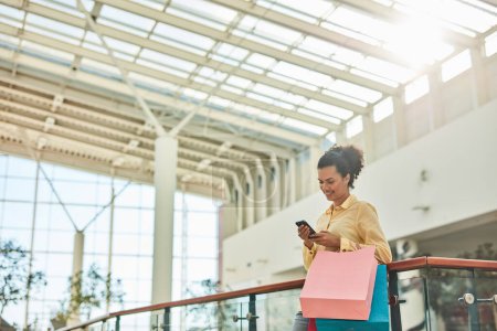 Téléchargez les photos : Une femme debout dans un centre commercial, tenant des sacs à provisions et un smartphone, souriant et communiquant avec les gens. - en image libre de droit