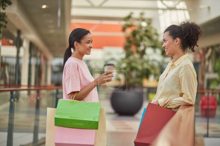 Foto de Un grupo de amigos disfrutando de un descuento de compras en el centro comercial, sonriendo y hablando junto con bolsas de compras. - Imagen libre de derechos