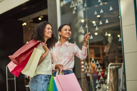 Téléchargez les photos : Deux femmes faisant leurs courses dans un centre commercial, pointant du doigt des articles en vente, souriant. - en image libre de droit