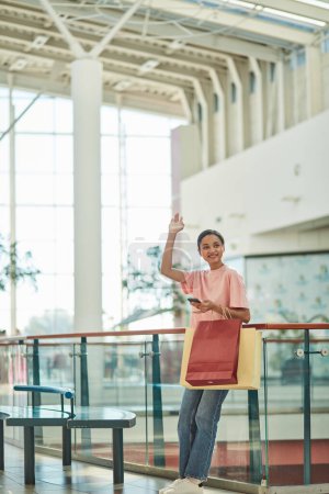 Foto de Una mujer sonriendo y saludando mientras está de pie con bolsas de compras en el centro comercial. - Imagen libre de derechos