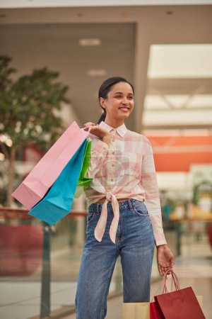 Foto de Una mujer caminando llevando bolsas de compras, disfrutando de una venta de descuento en una tienda de moda. Cliente feliz sonriendo, disfrutando de compras de estilo de vida. - Imagen libre de derechos