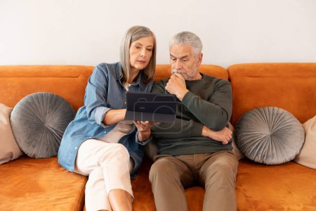 Téléchargez les photos : Femme est assise sur le canapé à la maison et montre quelque chose sur une tablette numérique à son mari. Un homme plus âgé regarde l'écran. - en image libre de droit
