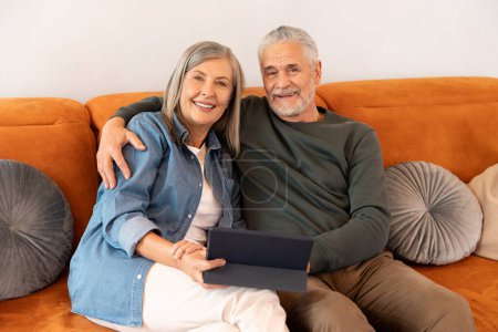 Photo for Portrait of smiling senior couple sitting in living room. Woman holding digital tablet with man on. sofa at home - Royalty Free Image