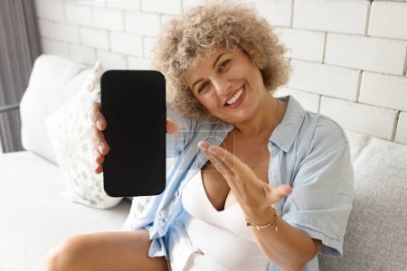 Téléchargez les photos : Joyeuse femme mature aux cheveux bouclés montrant un écran de smartphone vide, assise dans un environnement confortable à la maison. - en image libre de droit