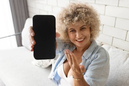 Téléchargez les photos : Femme aux cheveux bouclés joyeux montrant un écran de smartphone vierge, invitant à une expérience interactive. - en image libre de droit