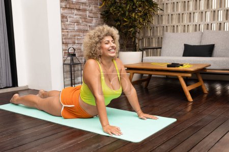 Téléchargez les photos : Femme aux cheveux bouclés joyeuse pratique le yoga sur un tapis à l'extérieur, rayonnant de positivité et de bien-être dans un cadre confortable à la maison. - en image libre de droit