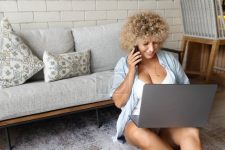 A cheerful young woman with curly hair multitasks with a laptop and phone, exuding a relaxed and productive vibe from her cozy home environment.