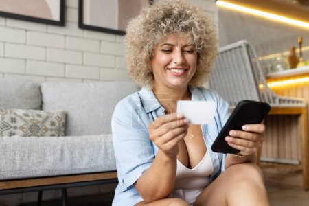 Portrait of a cheerful young curly-haired woman sitting on a sofa, smiling as she holds a smartphone and card.