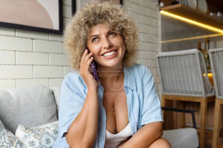 Photo for Cheerful young woman with curly hair having a pleasant phone call, sitting comfortably in a casual indoor setting. - Royalty Free Image