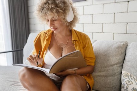 A focused woman with curly hair and headphones jotting down notes in a notebook while sitting comfortably on a couch at home.