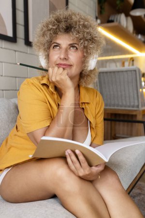 Photo for Curly-haired woman in contemplation, holding pencil, book, wearing headphones at home. - Royalty Free Image