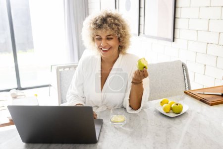 Foto de Una mujer rizada alegre está comiendo una manzana verde mientras usa una computadora portátil en una cocina brillante. Ella está vestida con un albornoz blanco, lo que sugiere un comienzo relajado, consciente de la salud de su día. - Imagen libre de derechos