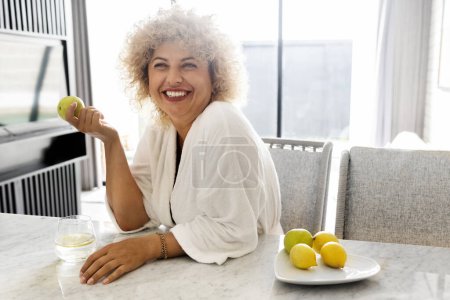Photo for Cheerful curly-haired mature woman holding an apple sitting in a sunlit kitchen. A moment of happiness and healthy lifestyle captured with a fresh and cozy home atmosphere. - Royalty Free Image