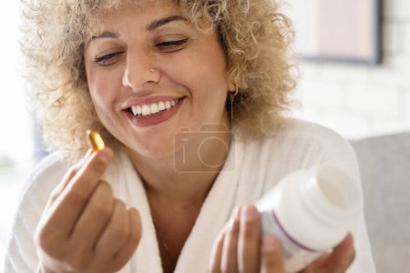 Foto de Rutina de bienestar matutino. Mujer feliz vestida de blanco tomando la cápsula de vitamina en casa. Ella sostiene una botella, teniendo en cuenta su rutina diaria de salud en un ambiente hogareño brillante y acogedor. - Imagen libre de derechos