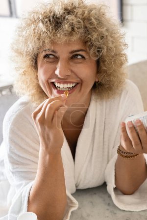 Foto de Retrato de una mujer feliz de pelo rizado con una bata blanca y sonriendo mientras toma una cápsula vitamínica. El concepto de salud, bienestar y suplementos diarios en un ambiente hogareño casual. - Imagen libre de derechos