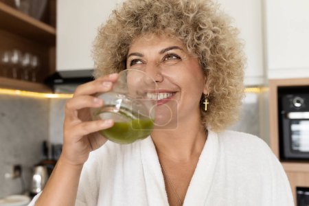 Photo for Joyful mature woman with curly hair drinking a fresh green smoothie in a bright kitchen, promoting a healthy lifestyle. - Royalty Free Image