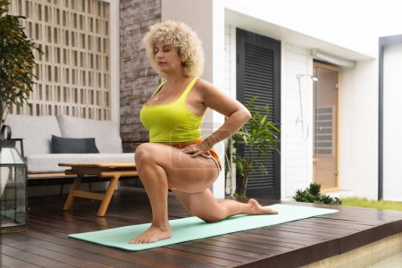 Téléchargez les photos : Une jeune femme aux cheveux bouclés frappe une posture de yoga sur un tapis à l'extérieur, incarnant une routine de fitness matinale saine dans un cadre serein. - en image libre de droit