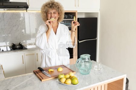 Foto de Mujer adulta joven vibrante en un albornoz comiendo limón fresco, preparando jugo de desintoxicación como parte de su rutina saludable de la mañana en un ambiente de cocina en casa brillante. - Imagen libre de derechos