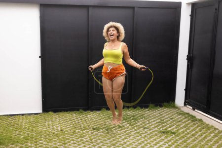 An energetic young adult woman smiles while jump roping outdoors, incorporating fitness into her daily morning ritual focused on health and vitality.