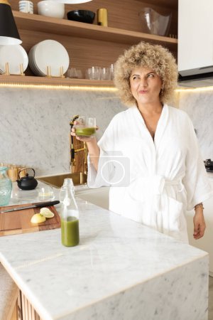 Téléchargez les photos : Femme souriante en robe blanche tenant un verre de jus vert dans une cuisine moderne, dépeignant un mode de vie sain, une routine matinale et des concepts de bien-être avec une touche de luxe. - en image libre de droit